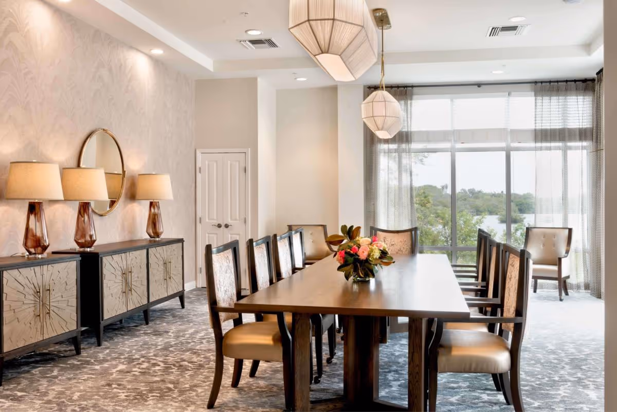 A bright and elegant dining room with a long wooden table surrounded by upholstered chairs. The room features large windows with sheer curtains allowing natural light and a view of greenery outside. On one side, there are two decorative sideboards with three table lamps and a round mirror above them. Two pendant lights hang from the ceiling above the table.