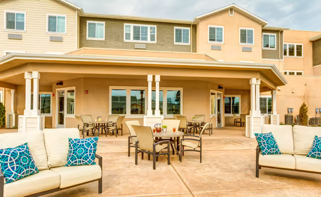 Outdoor patio area of a senior living facility with cushioned seating and tables with chairs arranged on a concrete surface. The building has multiple windows and a covered porch area.