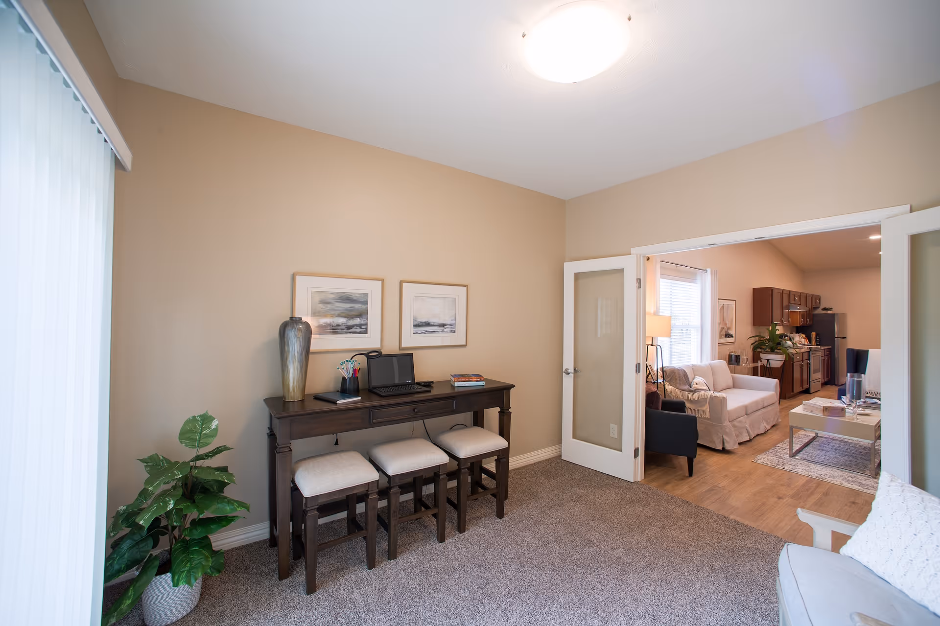 Interior view of a furnished apartment with a console table and stools in the foreground and a living room with sofa and kitchenette visible through open French doors.
