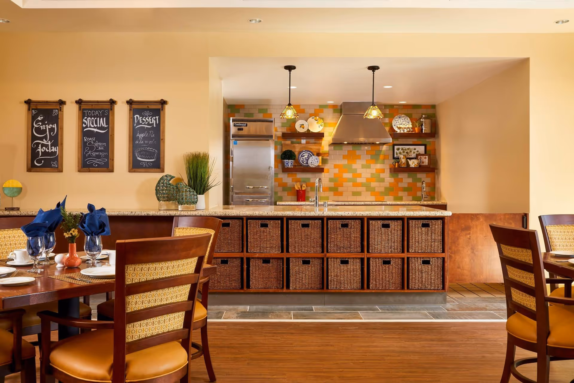 Dining room with set tables and chairs facing a serving counter with wicker storage baskets and a colorful tiled backsplash kitchen.