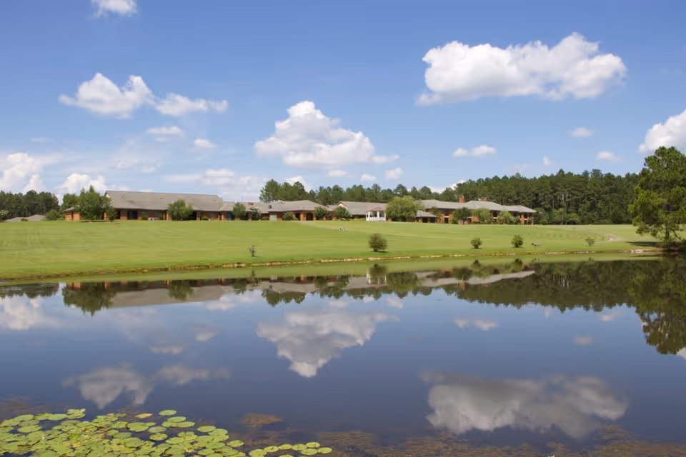 A large pond with lily pads in the foreground reflecting a partly cloudy blue sky. Beyond the pond is a wide expanse of green grass leading up to a single-story building complex surrounded by trees.