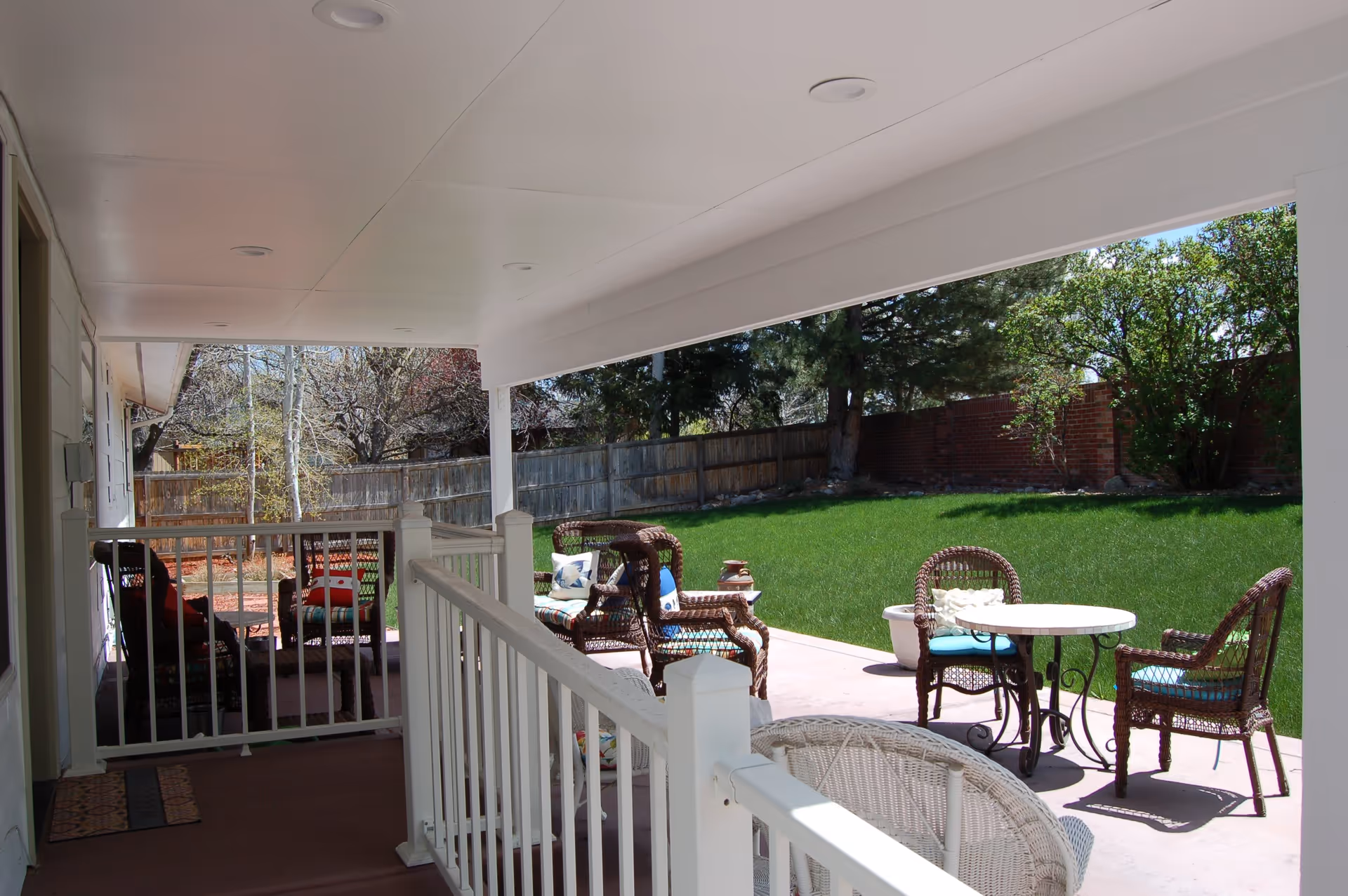 Covered porch with white railing and wicker patio furniture overlooking a green backyard lawn.