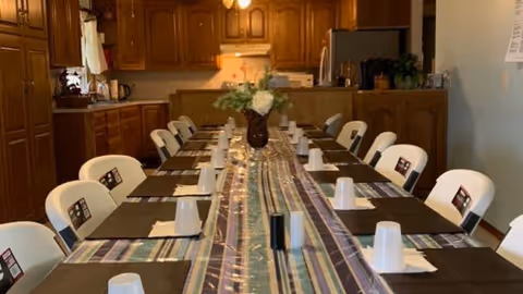 A long dining table set for a meal with placemats, napkins, and upside-down cups, situated in a kitchen area with wooden cabinets and appliances in the background. A vase with flowers is placed in the center of the table.