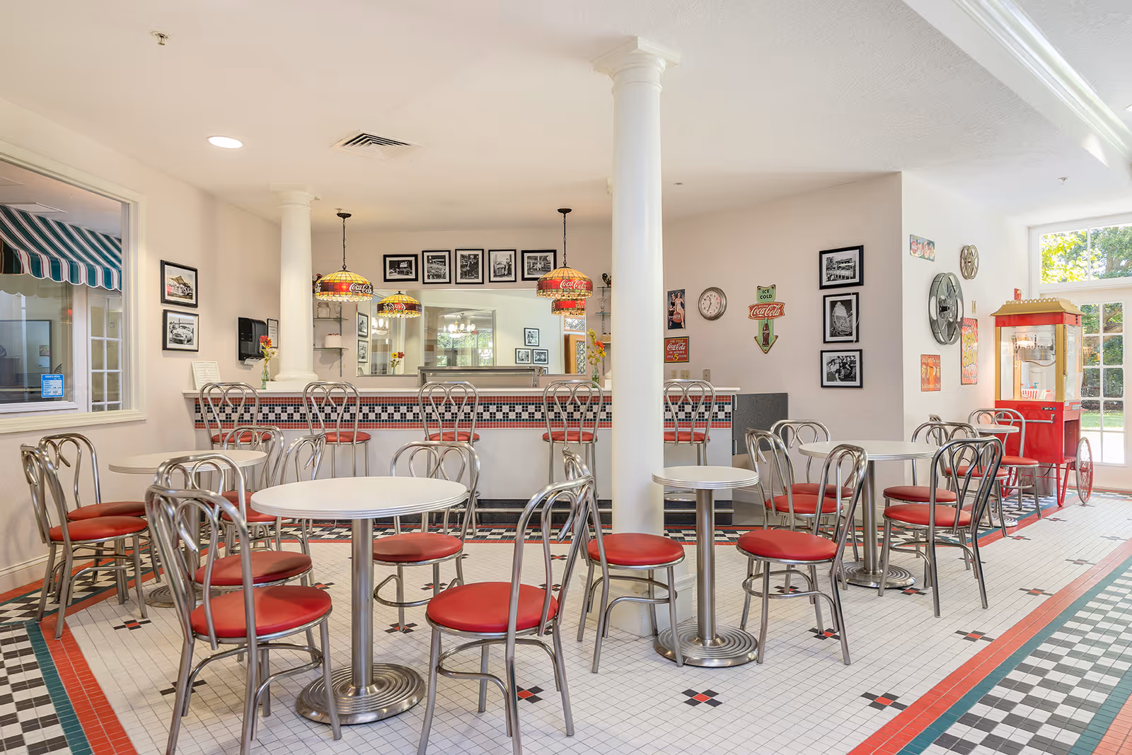Retro-style dining area with round white tables, red-cushioned chrome chairs, and a diner counter in a bright common room.