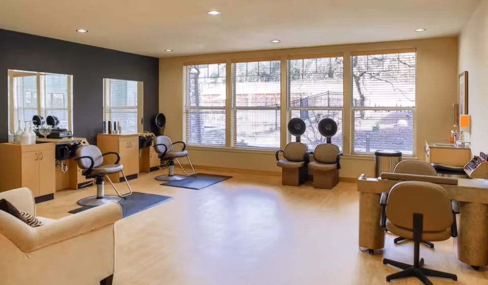 Interior view of a hair salon area in a senior living facility with beige walls and large windows. The room features salon chairs in front of mirrors, hair dryers, and a comfortable armchair. The floor is light wood, and the space is well-lit with recessed ceiling lights.