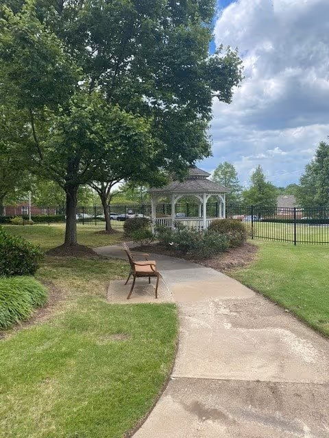A peaceful outdoor scene at Belmont Village Senior Living Memphis featuring a paved walkway leading to a white gazebo surrounded by green grass, trees, and shrubs. A wooden bench is placed along the path under a large tree, with a partly cloudy sky overhead.