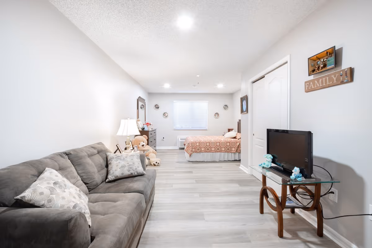 A bright and clean assisted living studio apartment featuring a gray couch with patterned pillows on the left, a small glass TV stand with a flat-screen TV on the right, and a bed with a patterned orange bedspread at the far end near a window with blinds. The room has light-colored wood flooring, white walls, and decorative items including a teddy bear, a lamp, and wall art with the word 'FAMILY'.
