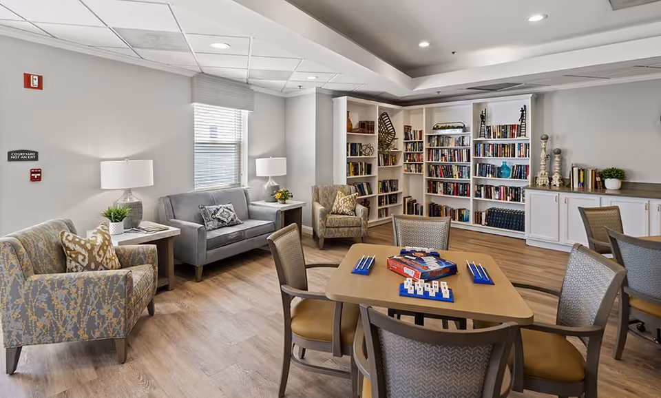 A cozy common area in a senior living facility featuring a wooden table with chairs around it and a board game set up on the table. The room has comfortable armchairs and a sofa with side tables and lamps. In the background, there are built-in bookshelves filled with books and decorative items. The floor is wooden, and the walls are painted light gray with a window letting in natural light.