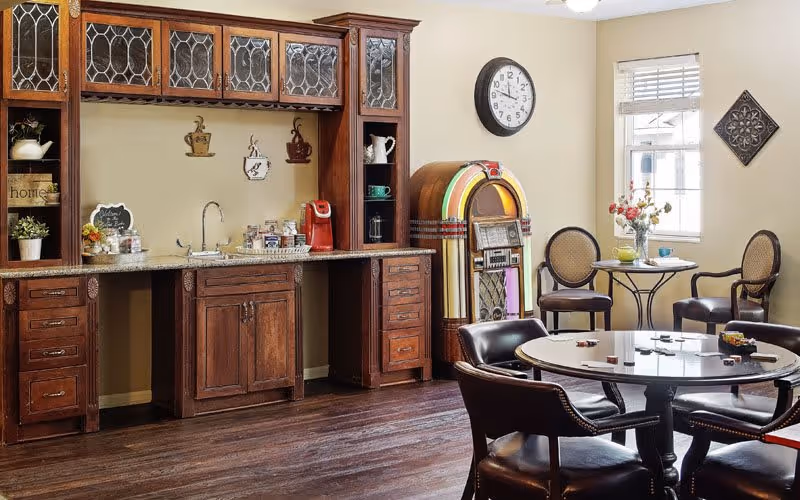 A cozy common area with a wooden cabinet featuring glass-paneled upper doors, a countertop with a sink, coffee maker, and various items. To the right, there is a vintage jukebox, a round table with four chairs, and a small table with two chairs near a window with blinds. The walls are decorated with a clock and a decorative wall piece.
