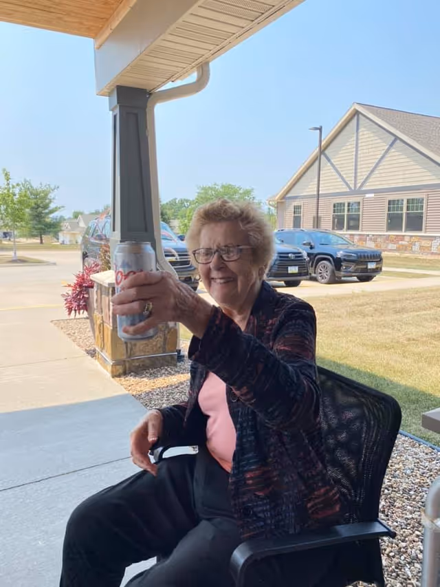 An elderly woman sitting outside on a black chair under a covered patio, smiling and holding up a can of Coors Light beer. Behind her, there are parked cars, a building with beige siding and stone accents, and a clear blue sky.