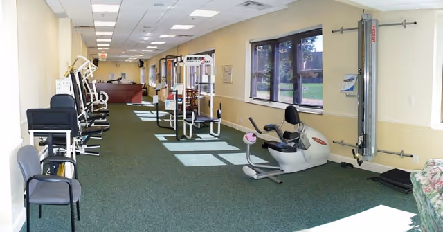 A long, narrow exercise room with various fitness equipment including stationary bikes, weight machines, and chairs along the left side. Large windows on the right side let in natural light, and the room has a green carpet and beige walls.