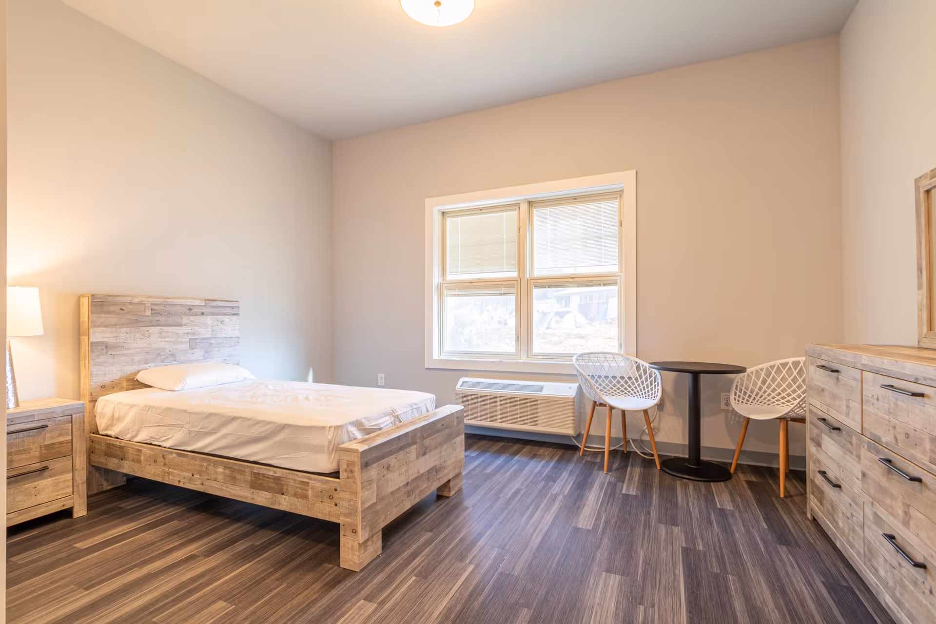 A bright, minimally furnished bedroom with a wooden bed frame and mattress, a wooden nightstand with a lamp, a window with blinds, two white modern chairs, a small round black table, and a wooden dresser with a mirror. The floor is dark wood laminate and the walls are light-colored.