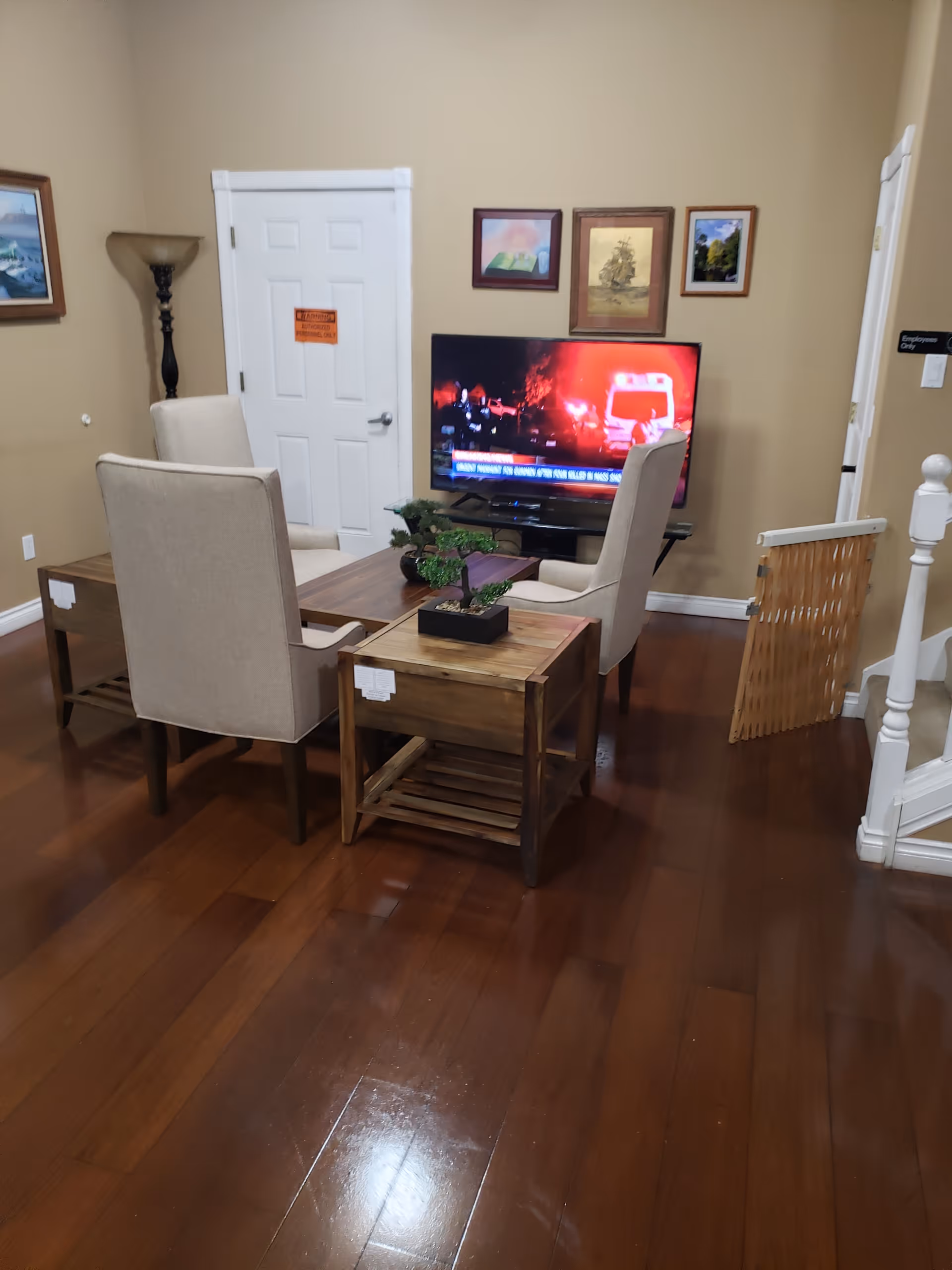 A cozy living room area with four beige upholstered chairs arranged around a wooden coffee table and side tables with small potted plants. A flat-screen TV is mounted on a stand against a beige wall, displaying a news broadcast. The room has wooden flooring, framed pictures on the wall, a floor lamp, and a white door with a sign. A baby gate is partially visible near a staircase.