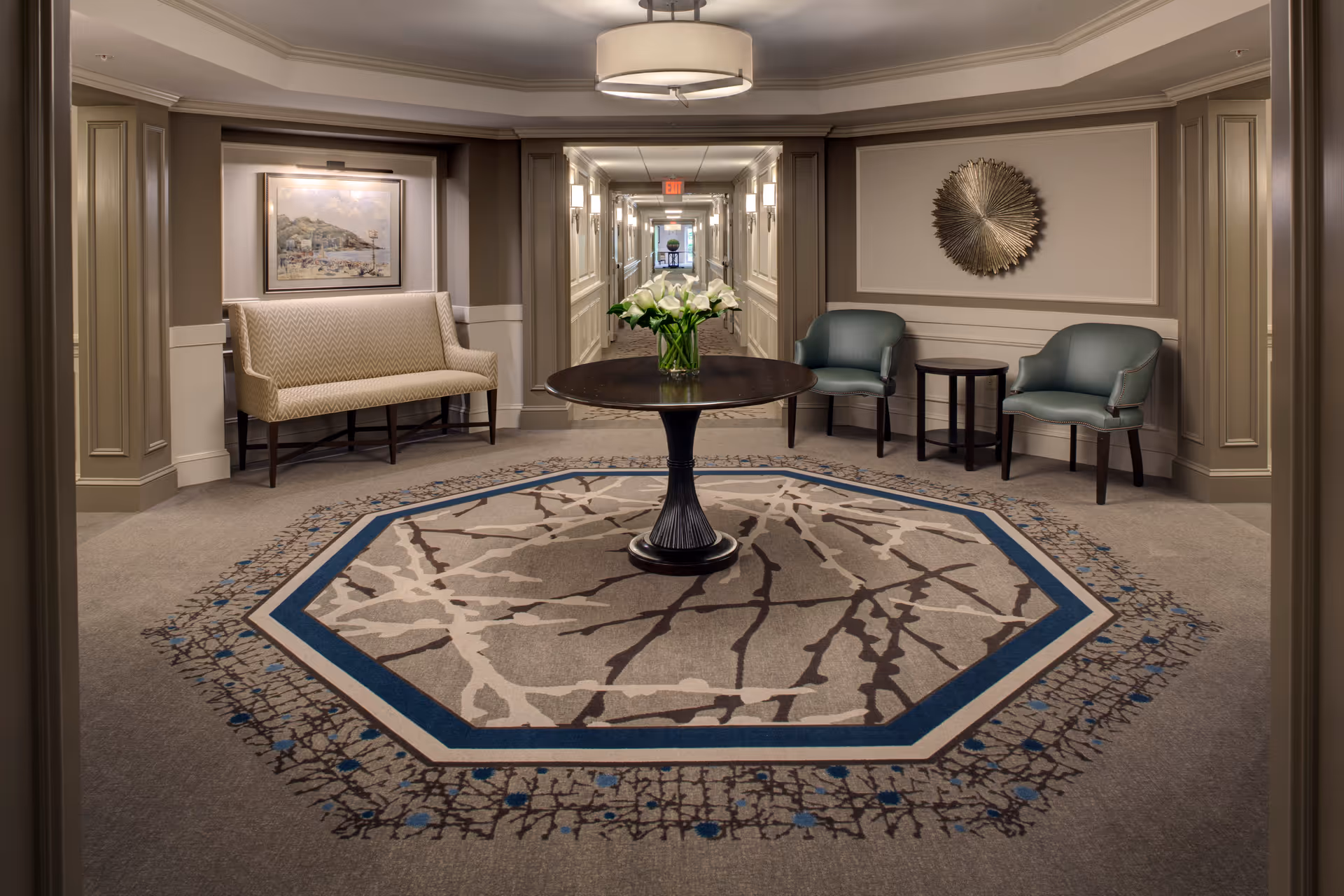 A well-lit hallway seating area with a round table holding a vase of white flowers in the center on a patterned octagonal rug. There is a beige cushioned bench with a framed painting above it on the left side, and two green armchairs with a small round table between them on the right side. The walls are decorated with molding and a metallic circular wall art piece. The hallway extends into the distance with multiple wall sconces and an exit sign visible.