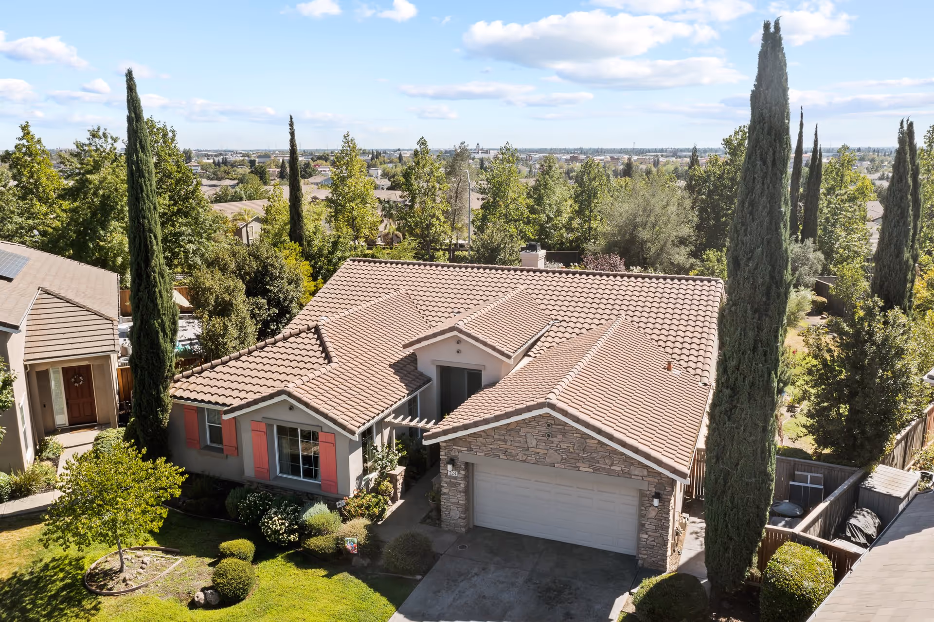 Aerial view of a single-story house with a tiled roof, stone facade on the garage, and red window shutters. The house is surrounded by tall trees and greenery, with a well-maintained lawn and bushes in the front yard. The sky is partly cloudy with blue patches.