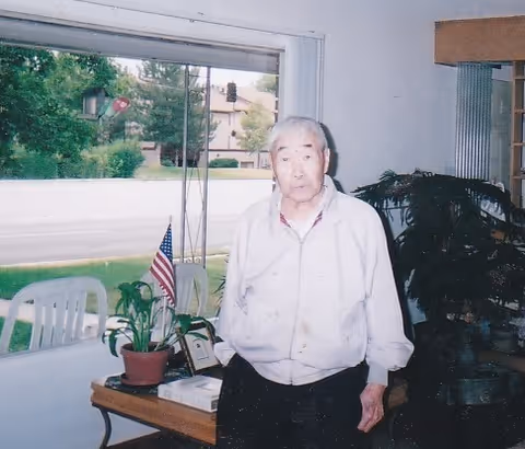 An elderly man standing inside a room near a large window with a view of a street and greenery outside. There is a small table with a potted plant, an American flag, and some framed items. The room has white walls and some plants in the background.