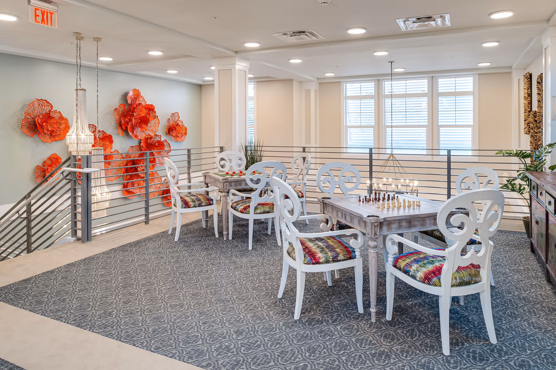 A bright, spacious common area with two wooden tables set up for games, one with a chessboard and the other with a checkers board. The tables are surrounded by white chairs with colorful patterned cushions. The room features a patterned carpet, large windows with white blinds, modern ceiling lights, and decorative red wall art resembling coral or flowers. A staircase with metal railings is visible on the left side.