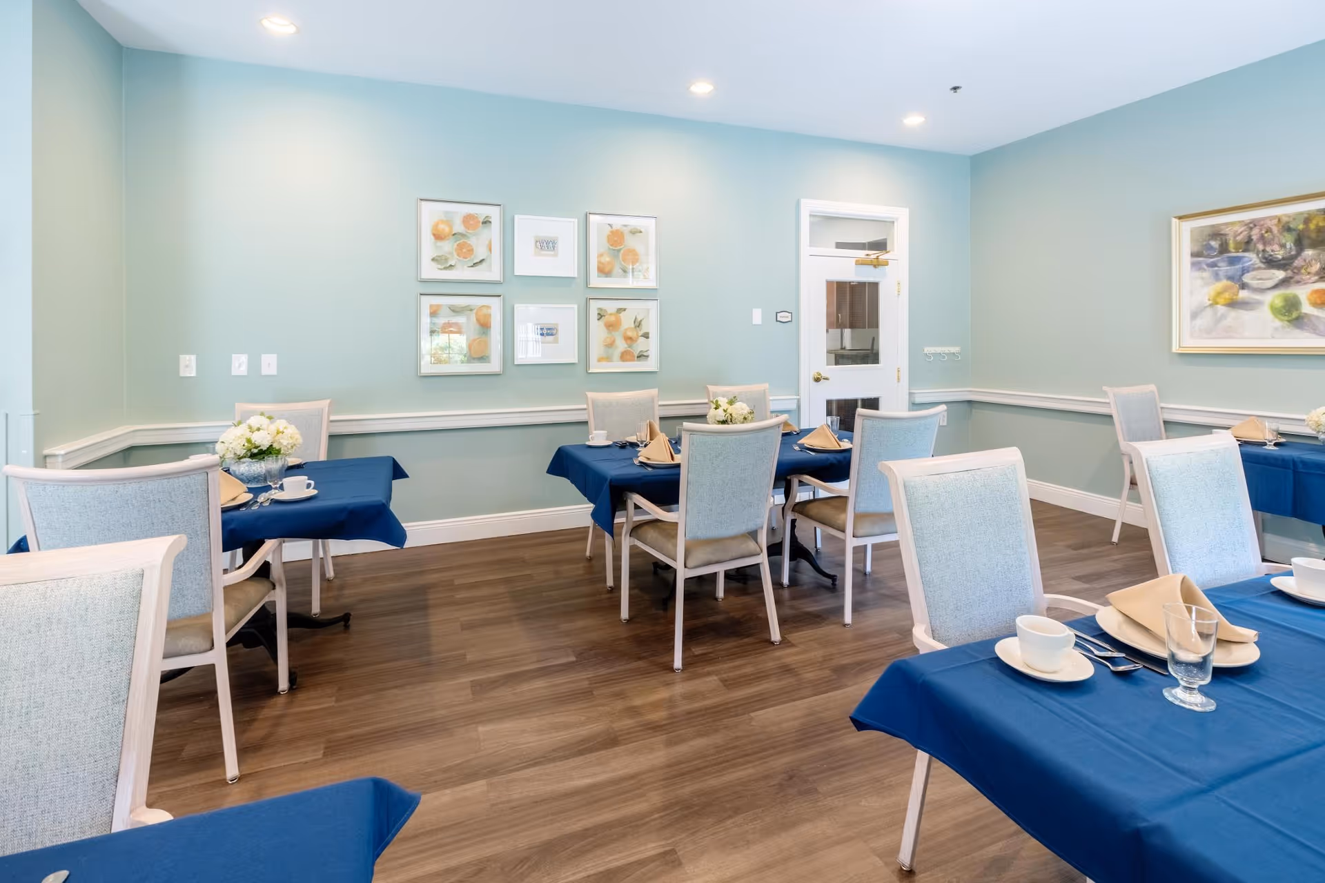 A dining room with several tables covered in blue tablecloths, each set with white plates, cups, glasses, and beige napkins. The room has light blue walls adorned with framed artwork and a wooden floor. Light-colored chairs surround the tables, and the room is well-lit with recessed ceiling lights.