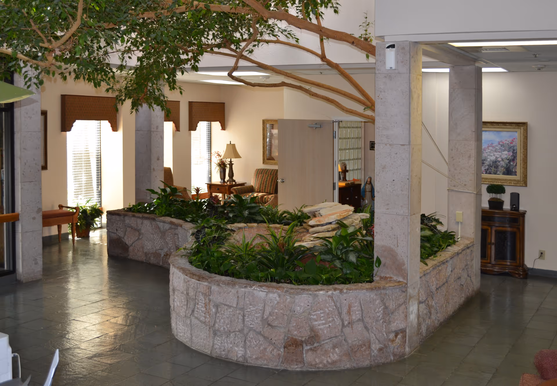 Interior lobby with a curved stone planter full of greenery and trees, columns, and a seating area with lamps and artwork.