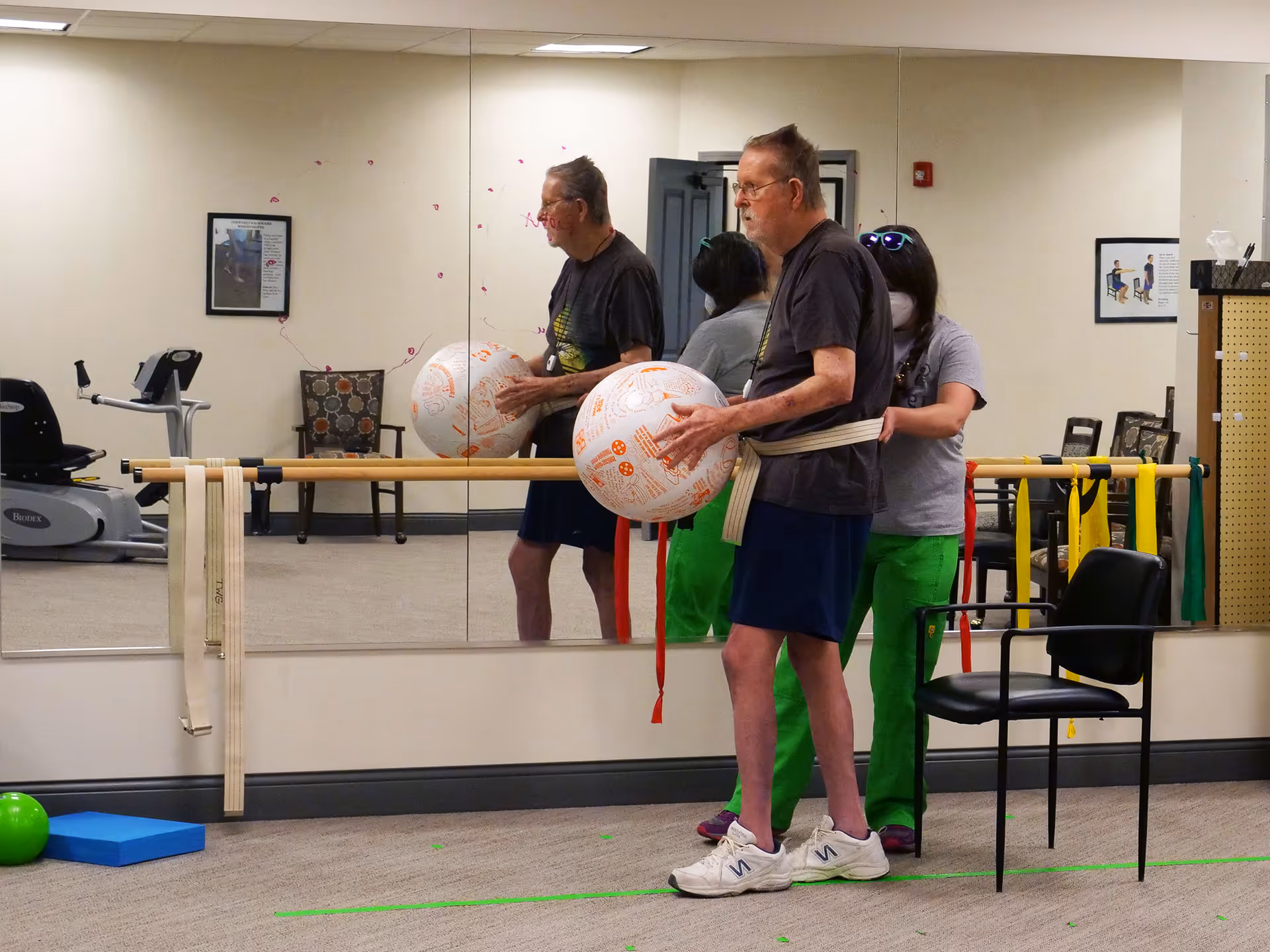 An elderly man holding a large exercise ball while a caregiver stands behind him in a room with a mirrored wall. The man is wearing a belt around his waist attached to the caregiver. The room has exercise equipment, chairs, and resistance bands hanging on a ballet barre.