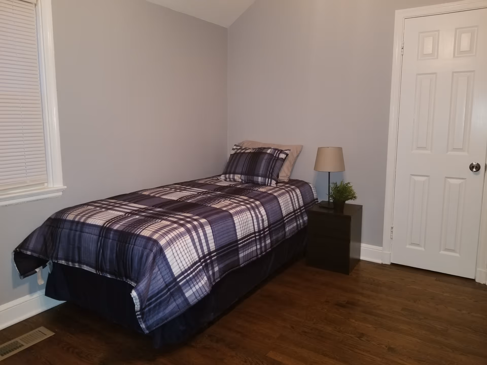 A simple bedroom with a single bed dressed in blue plaid bedding, a nightstand with a lamp and small plant, hardwood floor and a closed white door.