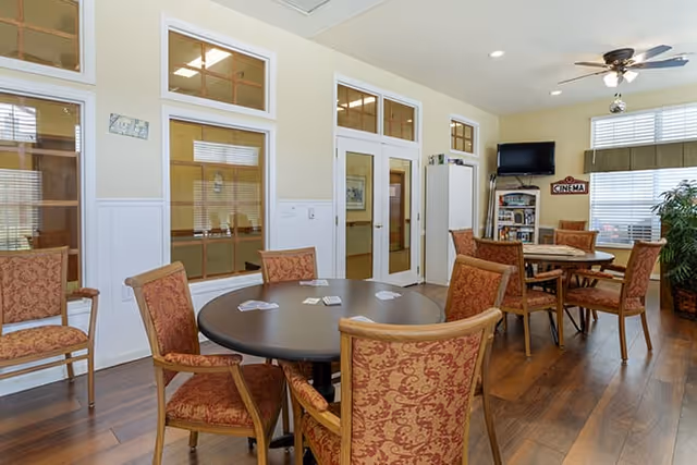A well-lit common area with round tables and upholstered chairs arranged for social activities. The room has wooden flooring, large windows with blinds, a wall-mounted TV, and a bookshelf. There is a sign on the wall that says 'CINEMA'.