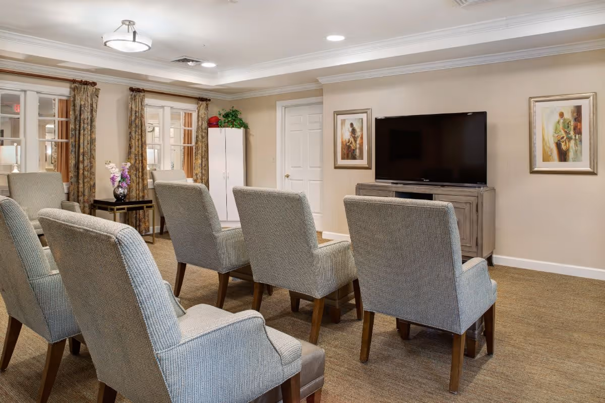 A cozy common area with six upholstered armchairs arranged in two rows facing a flat-screen TV mounted on a wooden cabinet. The room features beige walls, carpeted floor, two framed paintings on the wall, a white door, and windows with floral curtains. A small table with a vase of flowers is placed near the windows.