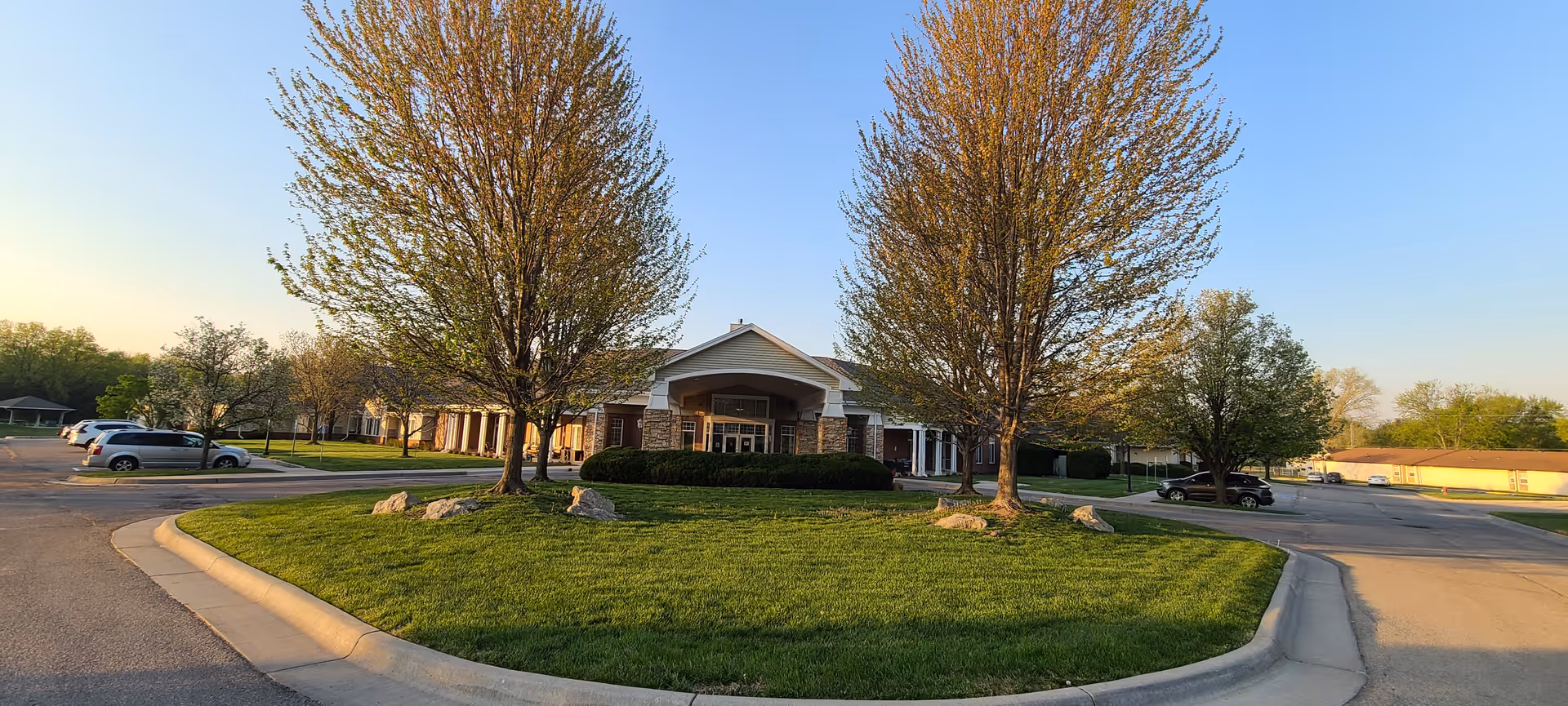 Front exterior view of Maria Court facility with a circular driveway, green lawn, and several trees. Cars are parked on both sides of the driveway under a clear blue sky.