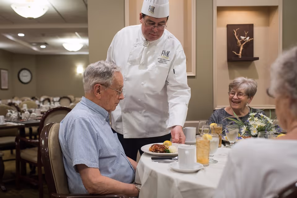 A chef in a white uniform and hat serves a plate of food to an elderly man seated at a dining table. Two elderly women are also seated at the table, one smiling and looking at the man. The table is set with glasses of iced tea, water, coffee cups, and a floral centerpiece. The background shows a dining room with multiple tables and chairs.