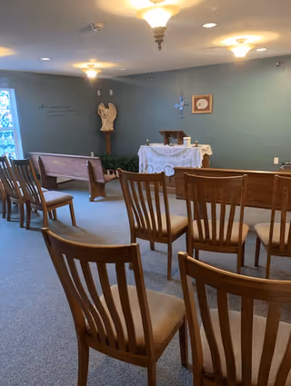 Interior of a small chapel or prayer room with wooden chairs arranged in rows facing an altar covered with a white cloth. The walls are painted light blue, and there is a statue of an angel, a cross, and framed artwork on the wall. Ceiling lights illuminate the room.