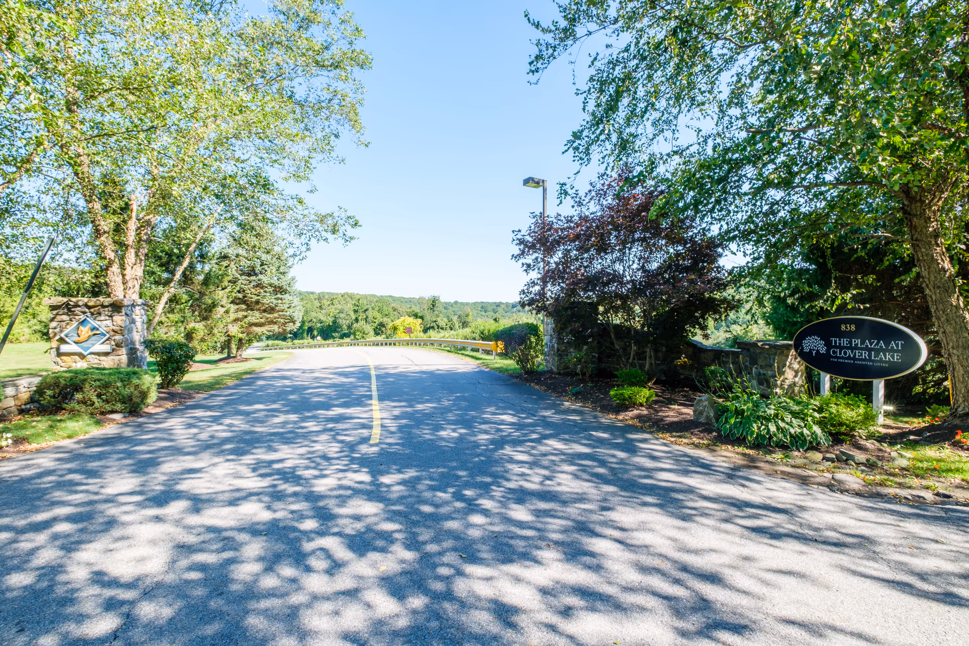 Entrance road to The Plaza at Clover Lake senior living facility, with trees and greenery on both sides and a clear blue sky overhead. There are two stone signposts on either side of the road, one with the facility name and logo.