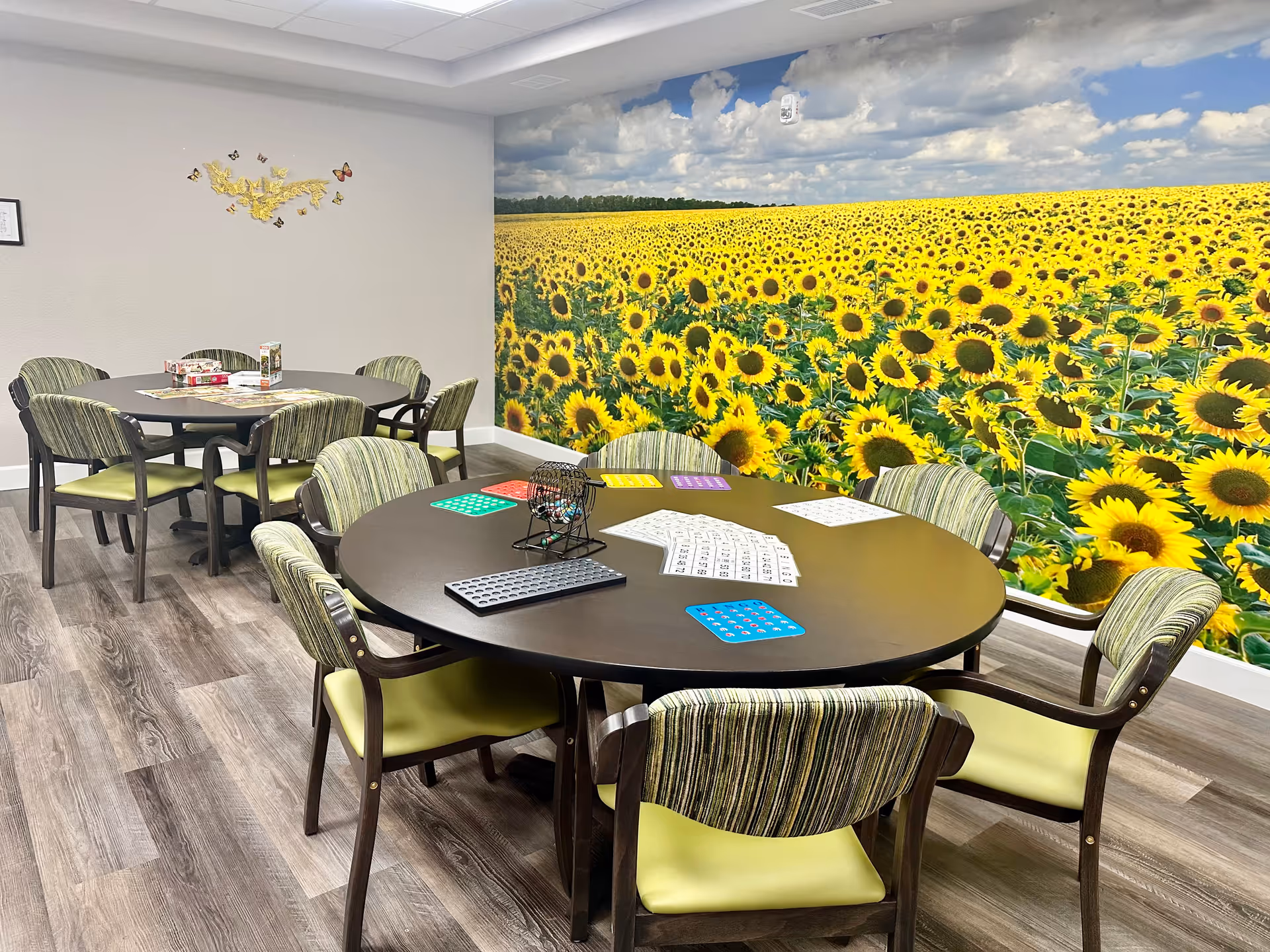 A bright activity room with two round tables surrounded by chairs with green cushions and striped backs. One table has bingo cards, a bingo cage, and other game materials on it. The room features a large wall mural of a sunflower field under a partly cloudy sky. The floor is wood-patterned, and there is a decorative butterfly wall art on the adjacent wall.