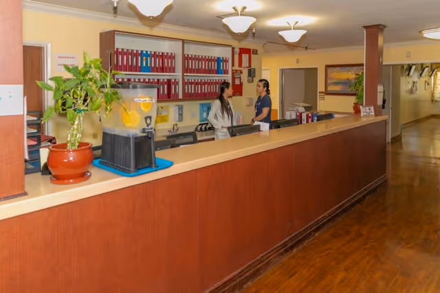 Reception area inside Atlantic Memorial Healthcare Center with a long wooden counter, two staff members talking behind the desk, a potted plant, a beverage dispenser with lemon water, and shelves with organized red binders on the wall. The hallway extends to the right with framed artwork and lighting fixtures on the ceiling.