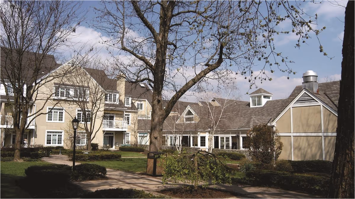 Multi-story senior living building front with a landscaped courtyard, trees, and walkways.