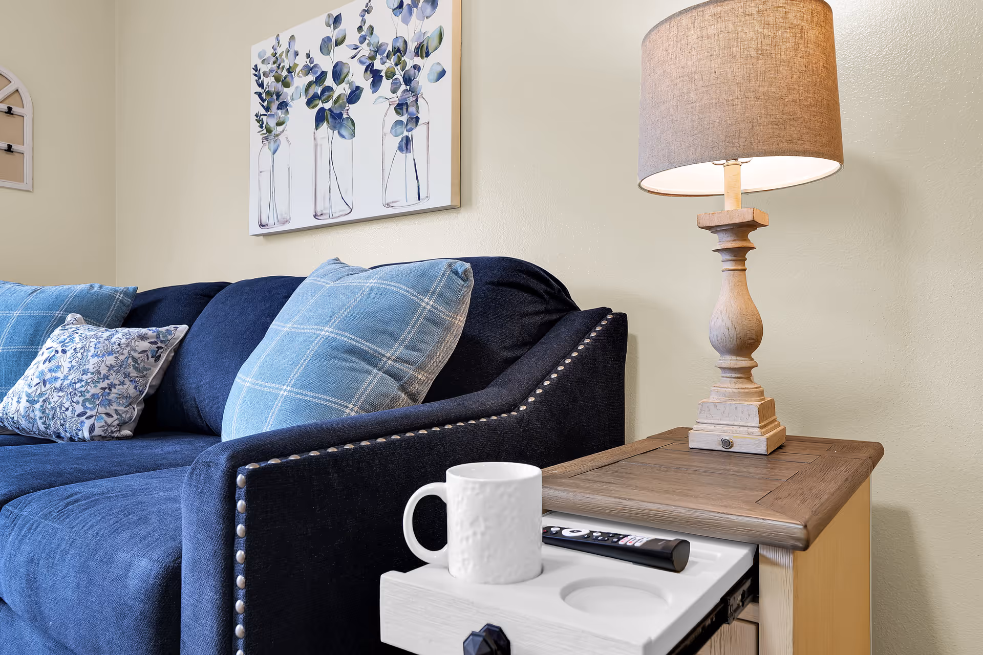 A cozy living room corner featuring a dark blue sofa with patterned blue and white cushions, a wooden side table with a beige lamp, a white mug, and a TV remote control. Above the sofa, there is a wall art painting of three glass jars with eucalyptus branches.