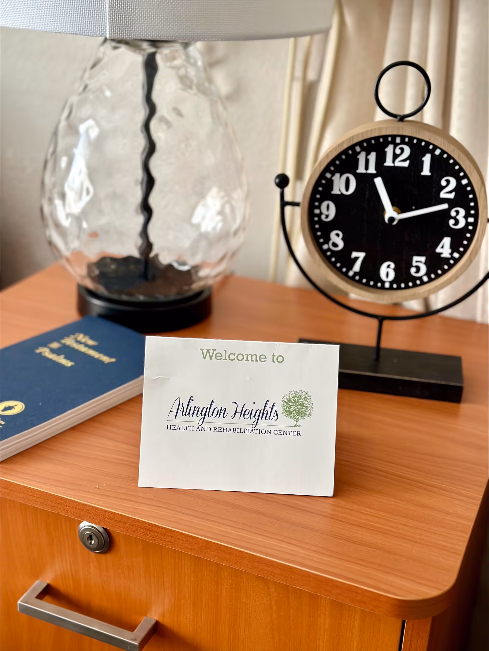 A wooden bedside table with a glass lamp, a black and wooden round clock showing the time as 11:12, and a welcome card that reads 'Welcome to Arlington Heights Health and Rehabilitation Center'. There is also a blue book titled 'Statement of Patient Rights' on the table.