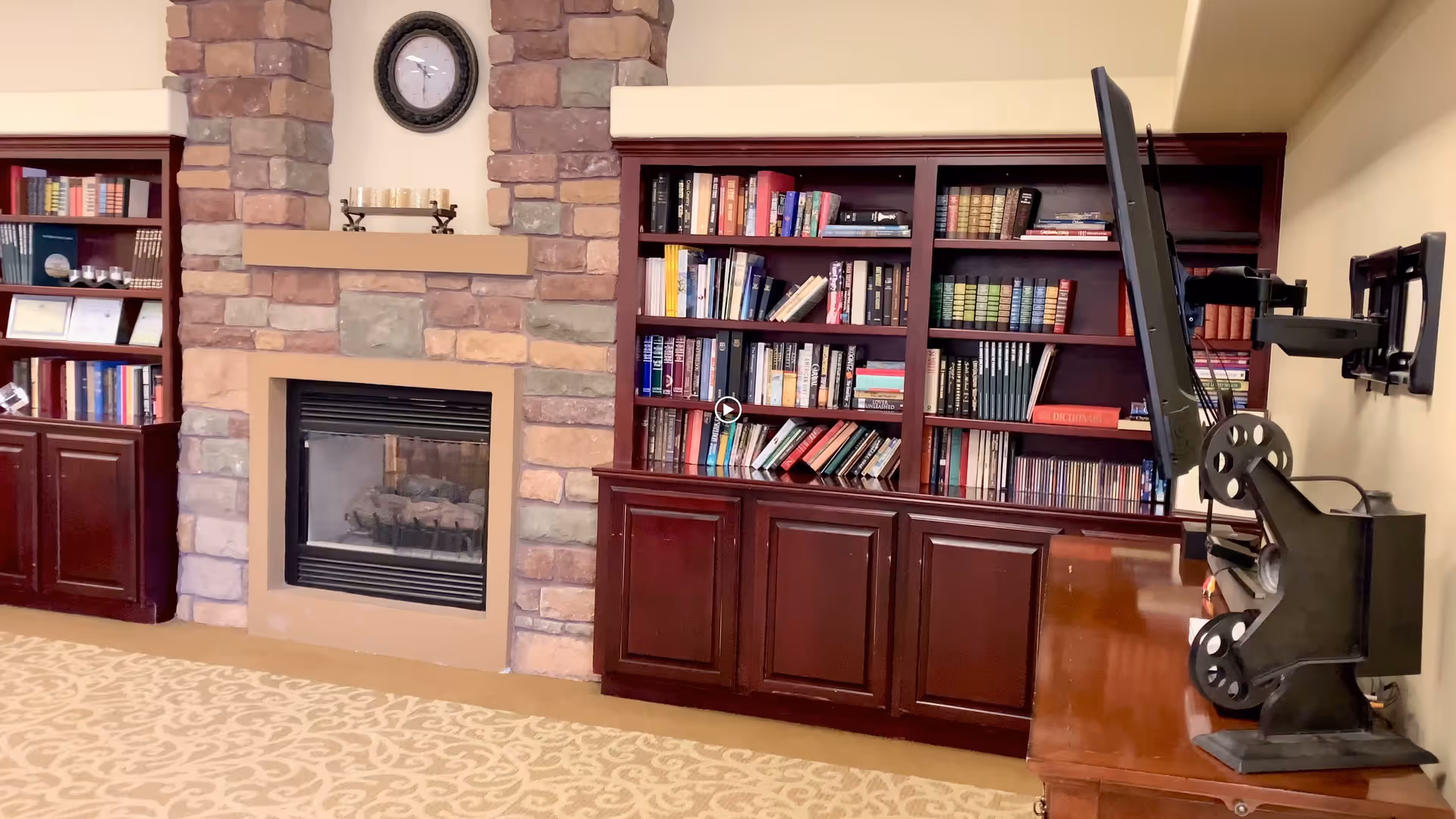 Interior view of a cozy living room area featuring a stone fireplace with a clock above it, flanked by dark wooden bookshelves filled with books. A flat-screen TV is mounted on the right wall with an adjustable arm, and a patterned carpet covers the floor.
