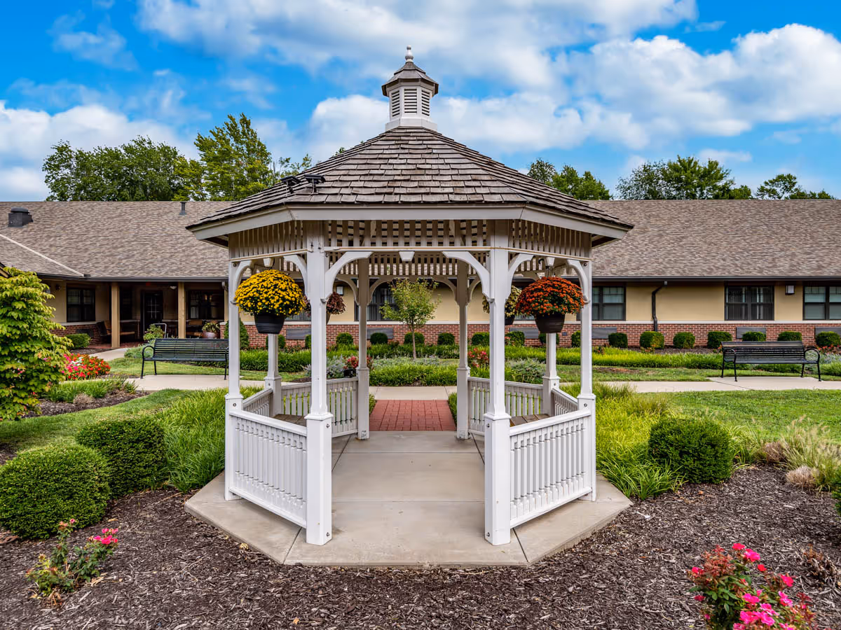 A white wooden gazebo with a shingled roof stands in the center of a well-maintained garden area with green bushes, colorful flowers, and paved walkways. In the background, there is a single-story building with beige walls, brick accents, and multiple windows. The sky is partly cloudy with blue patches.