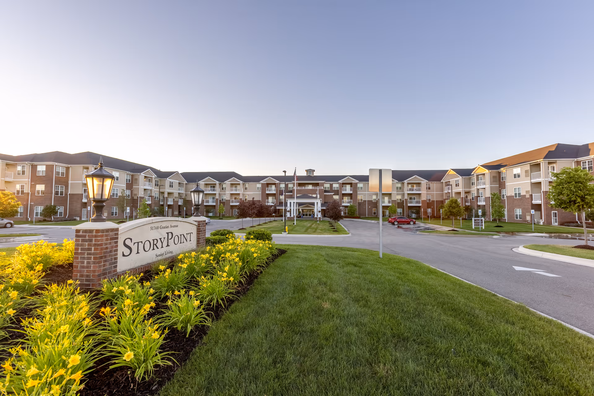 Wide exterior view of the StoryPoint Chesterfield senior living facility at sunset, showing a large three-story building with balconies, a well-maintained lawn, flower beds with yellow flowers, and a driveway leading to the entrance.
