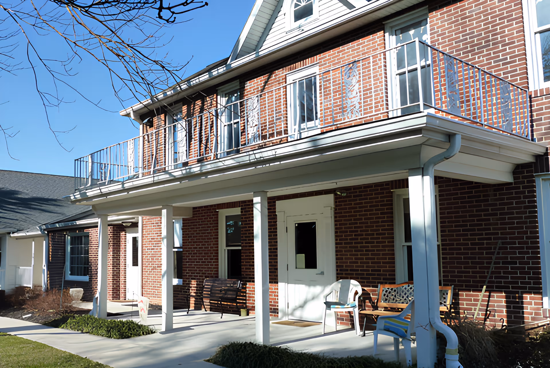 Exterior view of a brick building with a covered porch supported by white columns. The porch has several chairs and a bench, and there is a balcony with a metal railing above. The sky is clear and blue, and some bare tree branches are visible.