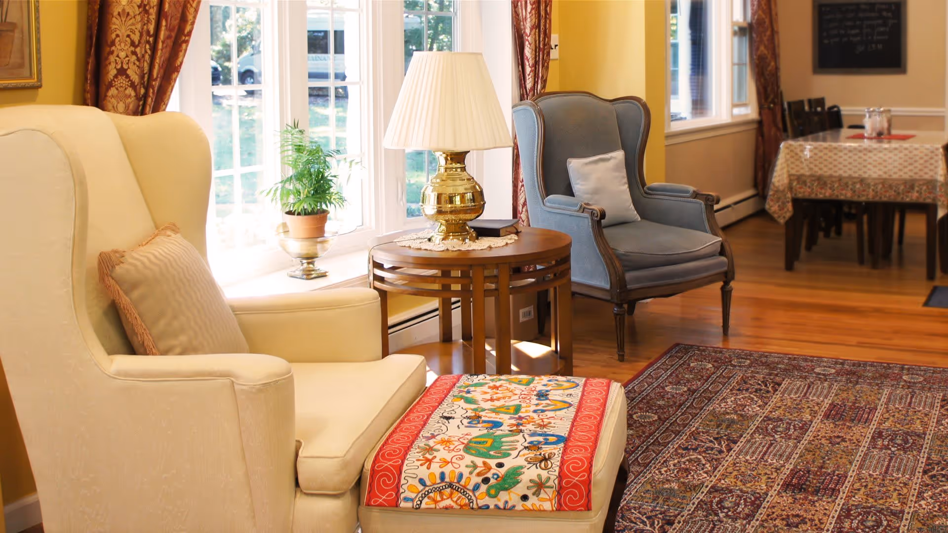 A cozy living area with a cream-colored armchair and matching ottoman featuring a colorful embroidered cover. Next to the armchair is a round wooden side table with a brass lamp and a small potted plant. In the background, there is a blue upholstered armchair and a dining table with a patterned tablecloth near large windows letting in natural light.