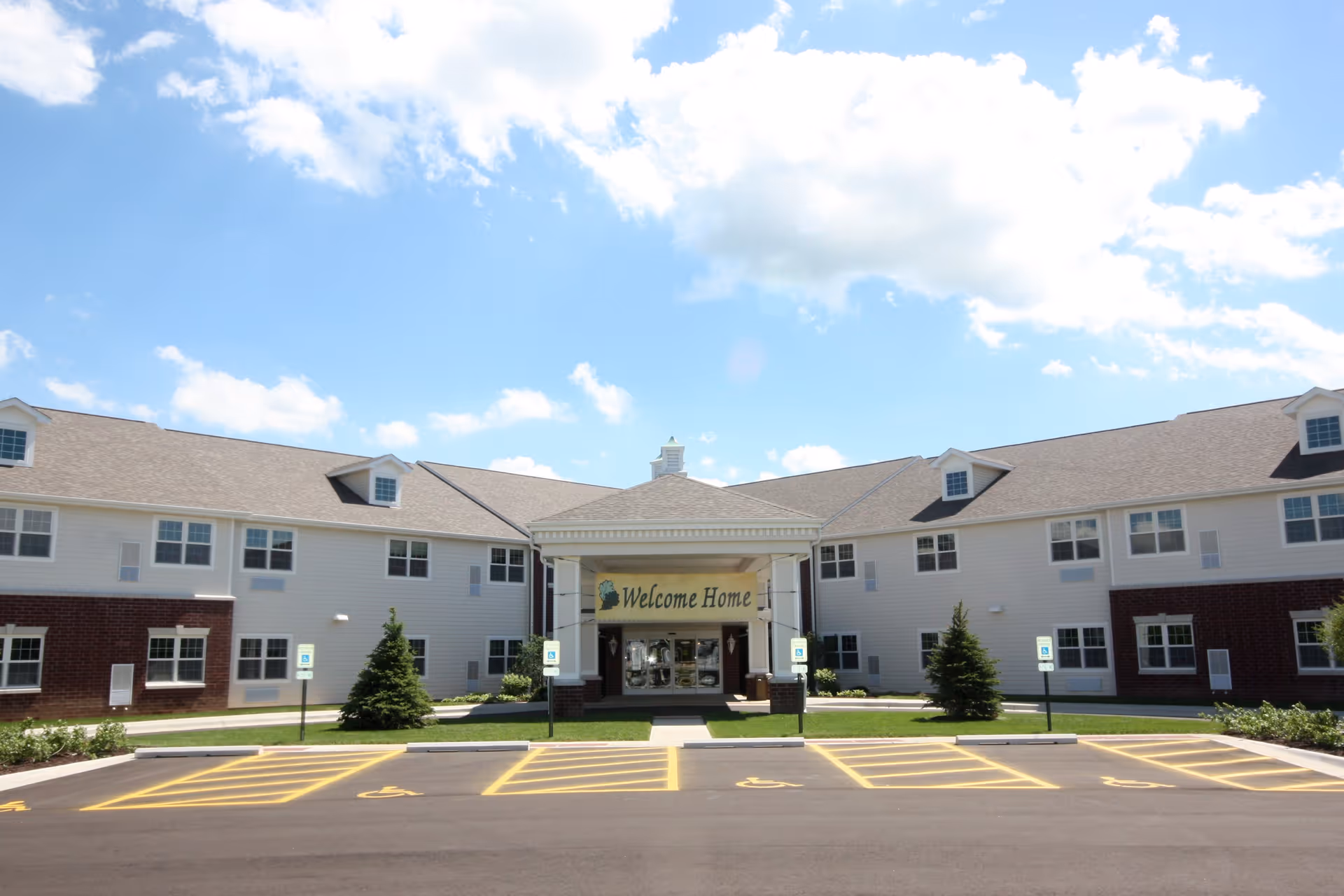 Front exterior view of a two-story senior living facility building with a covered entrance displaying a 'Welcome Home' sign. The building has white siding with red brick accents and multiple windows. There are several handicap parking spaces marked in yellow in front of the entrance. The sky is blue with scattered clouds.