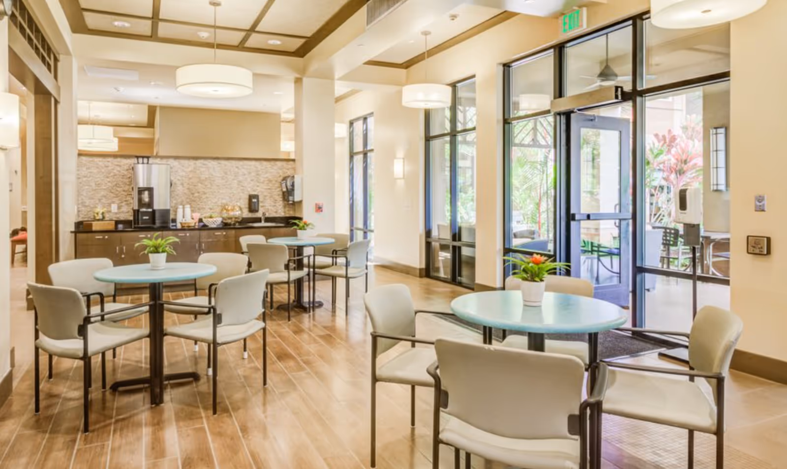 Sunlit communal dining area with round tables and chairs, a coffee station, and glass doors leading outside.