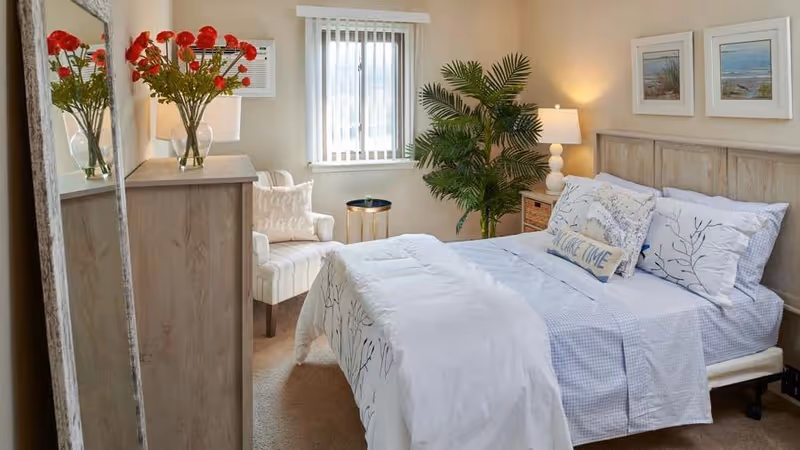 A cozy bedroom with a neatly made bed featuring white and blue bedding and decorative pillows. There is a wooden headboard with two framed pictures above it. To the right of the bed is a nightstand with a lamp and a large green potted plant. On the left side of the room, there is a wooden dresser with a vase of red flowers and a mirror leaning against the wall. A cushioned armchair with a pillow is positioned near a window with vertical blinds.