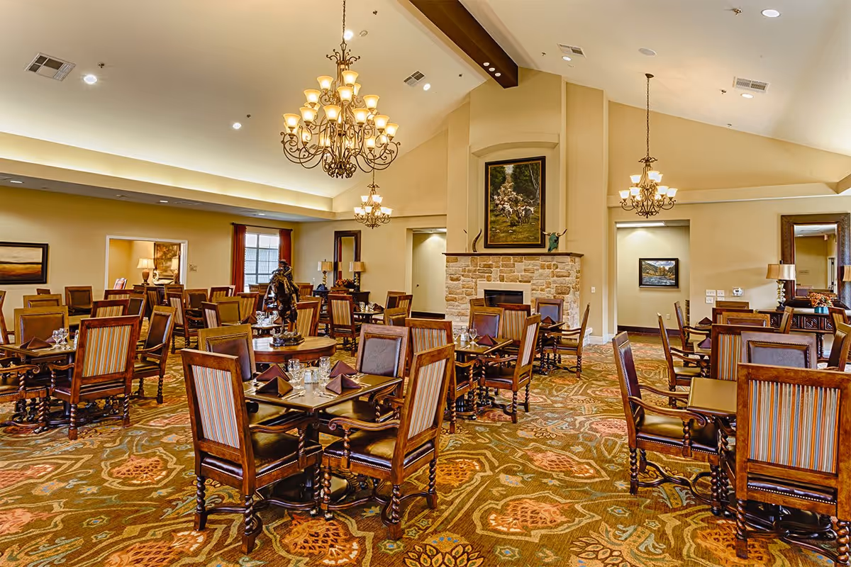 A spacious dining room with multiple wooden tables and chairs arranged neatly. The room features a patterned carpet, chandeliers hanging from a high ceiling, a stone fireplace with a painting above it, and warm lighting creating a cozy atmosphere.