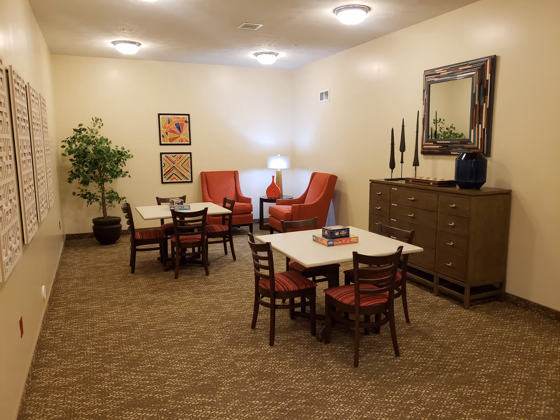 A cozy room with two tables each surrounded by four chairs with red and black striped cushions. In the corner, there are two red armchairs with a small table and a lamp between them. A large wooden dresser with a decorative mirror and three tall black candle holders is against the wall. The room has beige walls, carpeted floor, framed abstract art on the wall, and a potted plant in the corner.