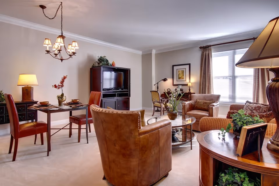 A cozy living room and dining area with beige walls and carpet. The living room features a brown leather armchair, a wicker armchair, a glass coffee table with a plant, and a TV cabinet with a flat-screen TV. The dining area has a small table set for two with red leather chairs and a decorative lamp on a side table. Large windows with curtains allow natural light to fill the space.