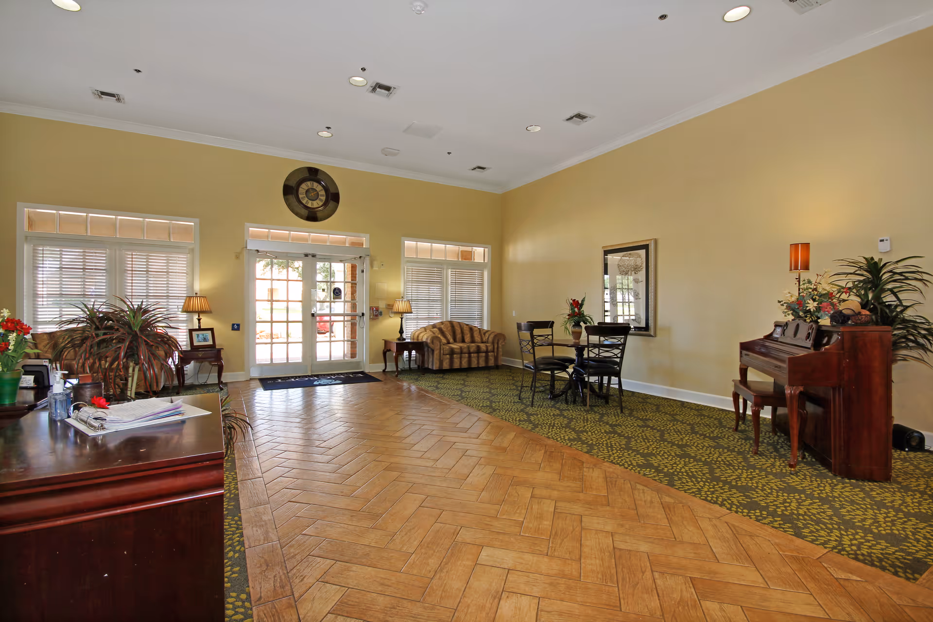 A spacious senior living facility common area with a wooden floor and patterned carpet. The room features a small round table with four chairs, a striped sofa, a piano with a stool, several plants, two table lamps, and large windows with blinds. Double glass doors lead outside, and a large clock is mounted above the doors.