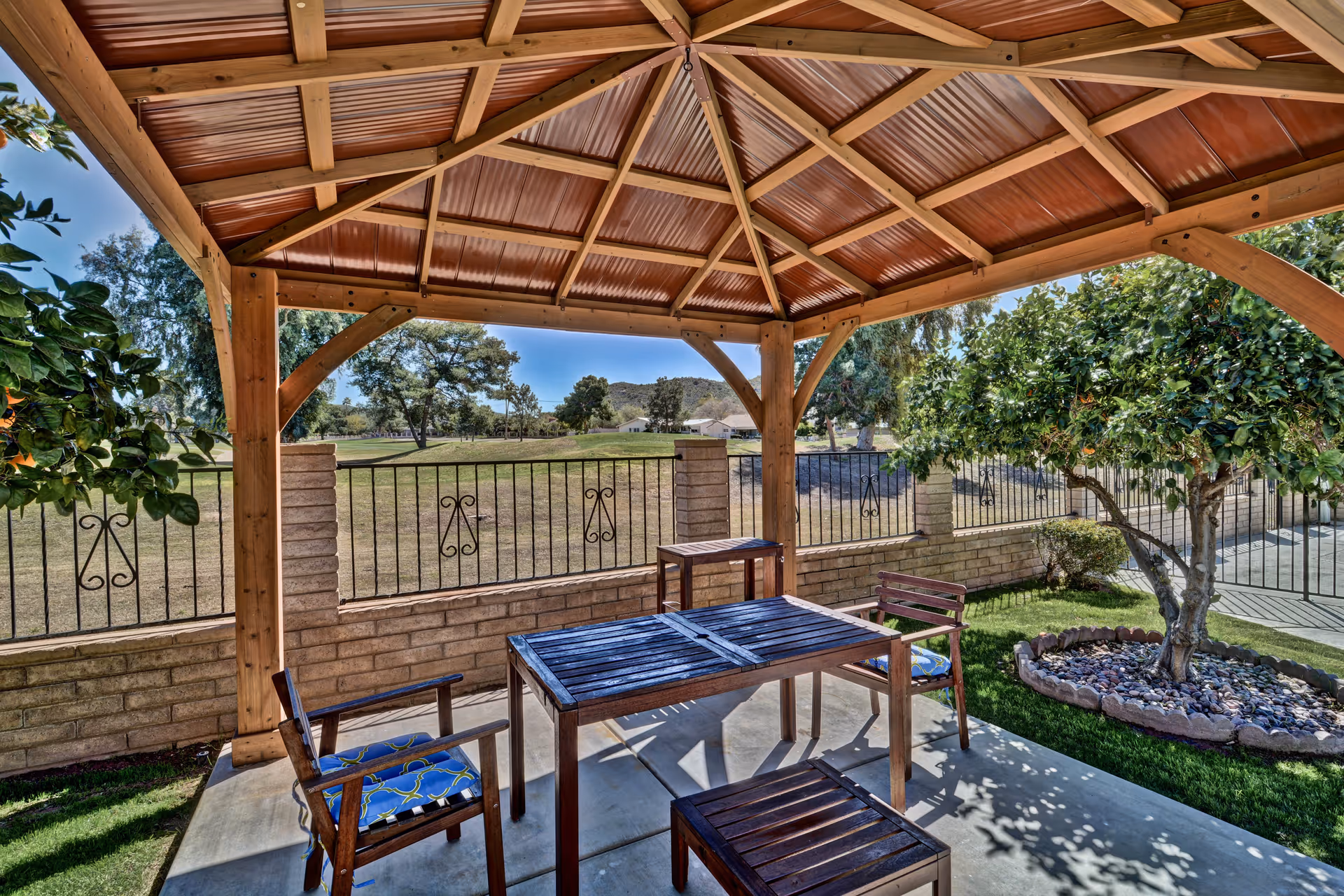Outdoor seating area under a wooden gazebo with a slanted roof, featuring a wooden table and four chairs with blue patterned cushions. The area is surrounded by a low brick wall with metal railings, and there are trees and a grassy lawn nearby under a clear blue sky.