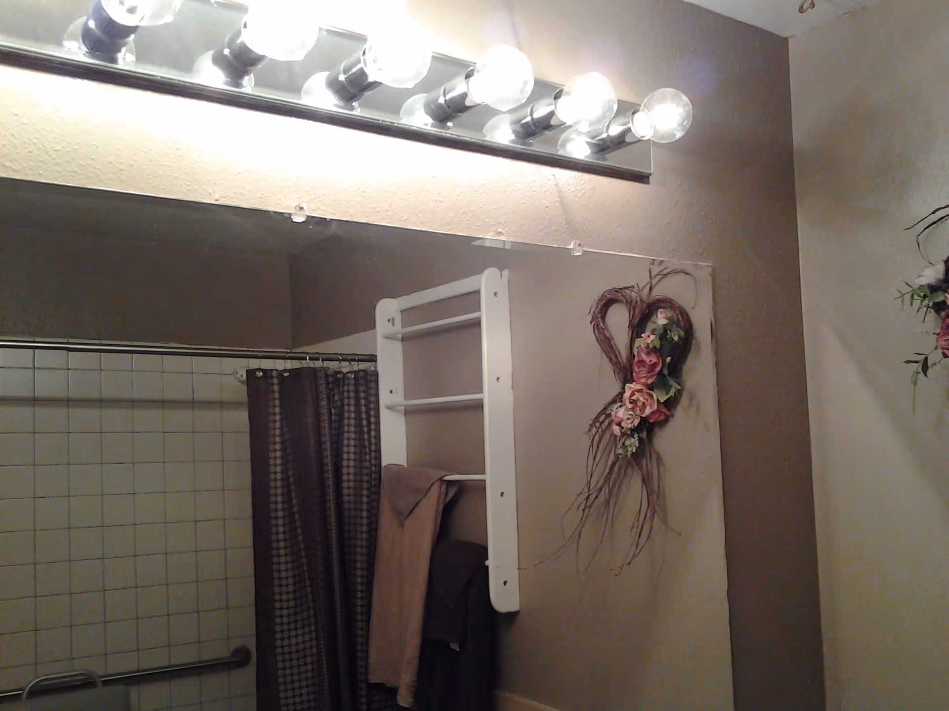 Bathroom interior with a large mirror reflecting a shower area with a brown patterned shower curtain and a grab bar. Above the mirror is a row of six round light bulbs. On the wall next to the mirror is a white towel rack holding two towels, and a decorative heart-shaped wreath with flowers is hanging on the wall.